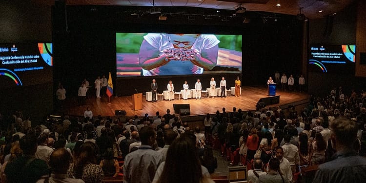 Panoramic view of the Second World Conference on Air Pollution and Health, with a lectern in the foreground and a large audience in the auditorium, where several flags and a screen presentation can be seen behind the speakers on stage.