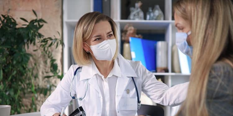 A female doctor wearing a mask, wearing a white coat and a stethoscope, is talking to a patient, also wearing a mask, in an office. The doctor is holding a pencil, and a table with documents and a plant can be seen in the background.