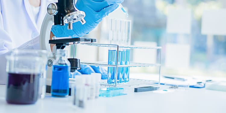 A scientist in a laboratory uses a pipette to add liquid to test tubes containing blue solutions, while a microscope and other laboratory instruments are visible on the table.