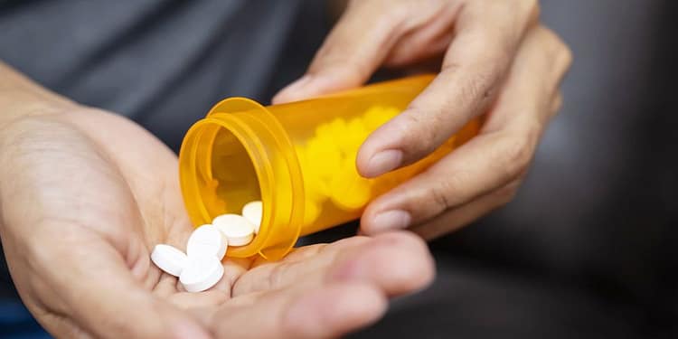 Una mano sostiene un frasco de medicamento naranja, mientras otra mano recibe pastillas blancas que caen del frasco en la palma.