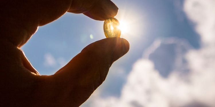 A hand holds a small transparent capsule against a background of blue sky with clouds, while sunlight shines through the capsule, creating a luminous effect.