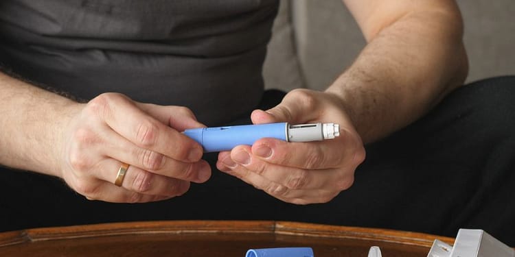Una persona sostiene un dispositivo de inyección azul en sus manos, con un cartucho adicional y varias agujas desechables sobre una mesa de madera.
