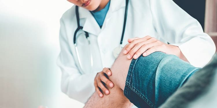 A doctor examines a patient's knee while they are both seated. The doctor is wearing a white coat and has a stethoscope around his neck.