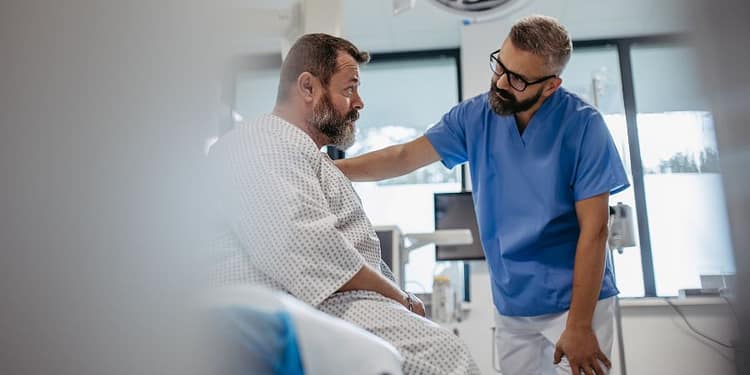 Un médico con gafas y camiseta azul se inclina hacia un paciente mayor con barba en una habitación de hospital, ambos mostrando expresiones serias. El paciente viste una bata de hospital y parece estar recibiendo atención médica.