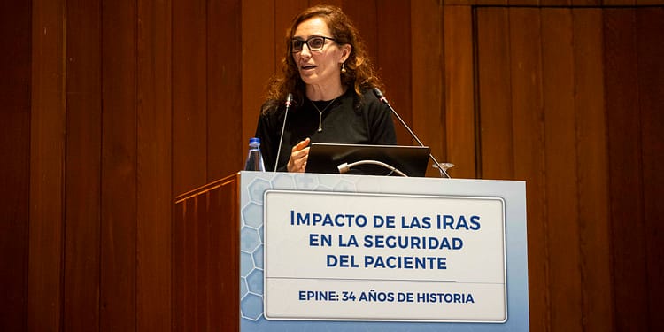 Mujer de cabello rizado y gafas, hablando en un podio con un letrero que dice "Impacto de las IRAS en la seguridad del paciente". Fondo de madera.