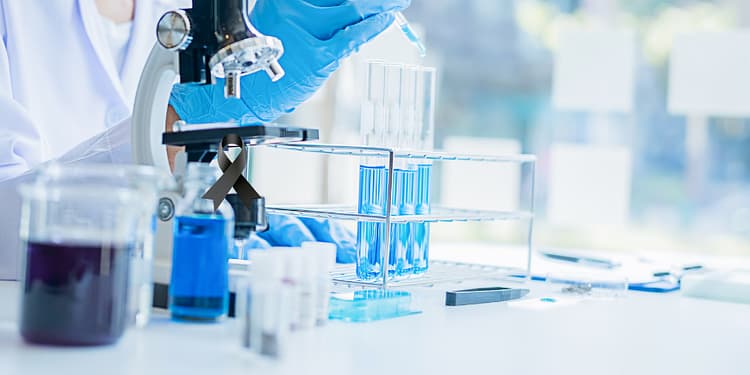 Mujer con guantes azules utilizando un microscopio y manipulando pipetas en un laboratorio, con tubos de ensayo y soluciones de diferentes colores sobre la mesa.