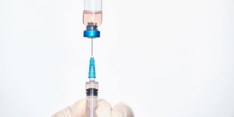 A gloved hand holding a syringe as it is filled with liquid from a clear vial containing a pink liquid.