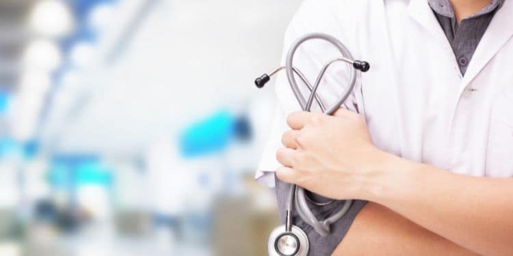 A healthcare professional in a white coat holds a stethoscope in his crossed arms, with a blurred background of a medical facility.
