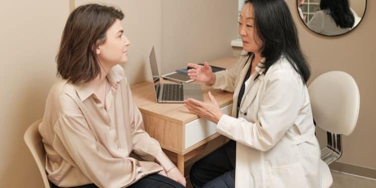 A woman with brown hair sits, listening attentively to a dark-skinned female doctor in a white coat, who is talking animatedly in front of a table with a laptop, in a modern office with beige walls and a circular mirror on the wall.