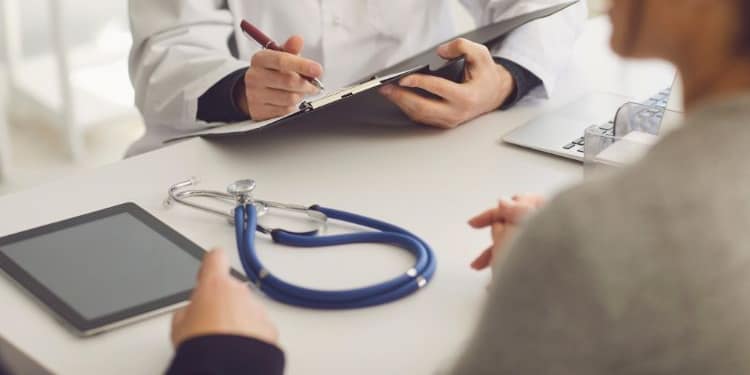Un médico, con una bata blanca y un bolígrafo en la mano, examina un documento mientras escucha a una paciente sentada frente a él. En la mesa hay un estetoscopio y una tableta.