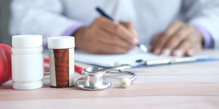 A doctor in a white coat writes on a sheet of paper while a stethoscope and medication bottles are visible on the table.
