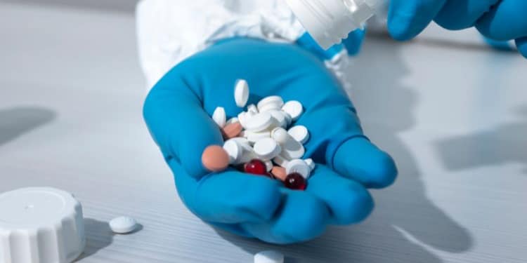 A blue-gloved hand holds several different colored tablets, while another hand pours more pills from a white bottle into the palm.