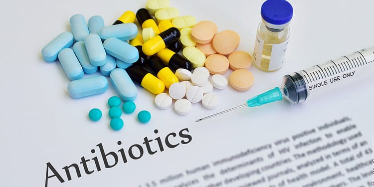 A variety of medications, including pills of different colors and shapes, a syringe and a bottle, on a document that says "Antibiotics."