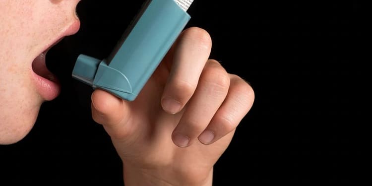 Person holding an asthma inhaler near his mouth, on black background.
