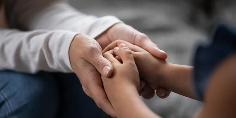 Una mano adulta sostiene suavemente las manos de un niño, ambas entrelazadas, en un ambiente acogedor.