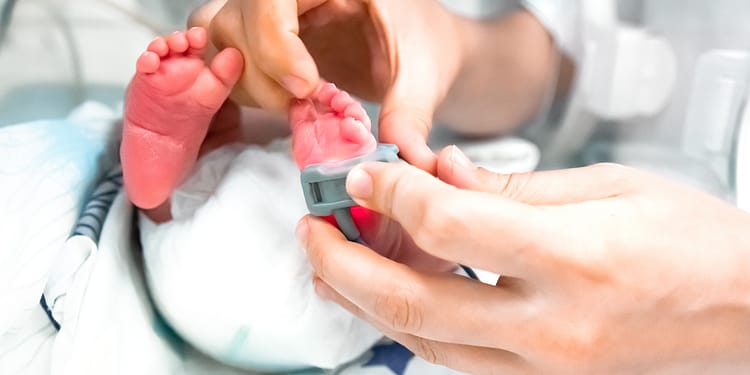 Un bebé con pies pequeños está siendo examinado por una mano que coloca un dispositivo de monitoreo en uno de sus pies, mientras descansa sobre una superficie equipada con sábanas médicas.