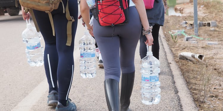 Group of people walking along a road, each carrying large bottles of water while wearing backpacks.