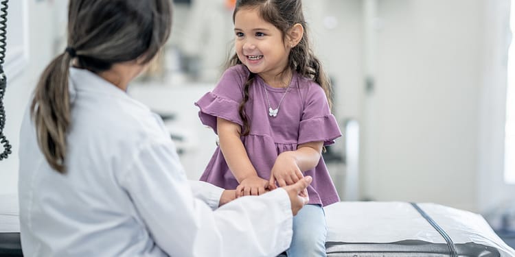 Smiling girl in purple blouse sitting on a medical examination table, holding the hands of a person with his back turned.