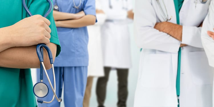 Group of health professionals, with a doctor in a white coat and others in colorful uniforms, some with stethoscopes.