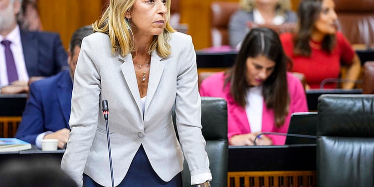 A woman standing in a parliament building, with long blonde hair, wearing a light-coloured jacket. In the background, other attendees and tables with computers.
