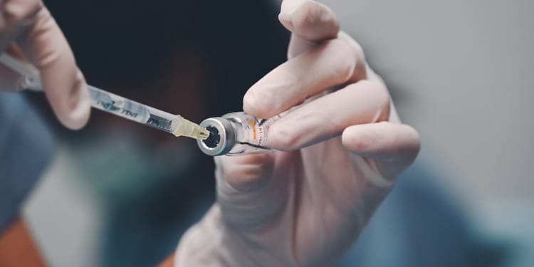 A gloved hand holds a syringe that is inserted into a vial, preparing to draw up a liquid.
