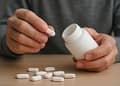 Una persona sostiene un frasco blanco de pastillas mientras se prepara para sacar una pastilla blanca, con más pastillas esparcidas sobre una superficie de madera.