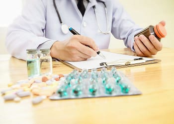 Un médico con un estetoscopio vestido con bata blanca sostiene un frasco de medicamento en una mano y escribe en un formulario con la otra, mientras hay viales y una variedad de pastillas sobre la mesa.