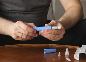 Una persona sostiene un dispositivo de inyección azul en sus manos, con un cartucho adicional y varias agujas desechables sobre una mesa de madera.