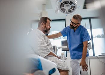 Un médico con gafas y camiseta azul se inclina hacia un paciente mayor con barba en una habitación de hospital, ambos mostrando expresiones serias. El paciente viste una bata de hospital y parece estar recibiendo atención médica.