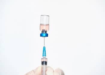 Una mano con guante sosteniendo una jeringa mientras se llena con líquido de un vial transparente que contiene un líquido rosado.