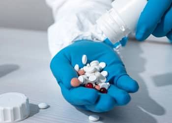 Una mano con guantes azules sostiene varias tabletas de diferentes colores, mientras otra mano vierte más pastillas de un frasco blanco en la palma.
